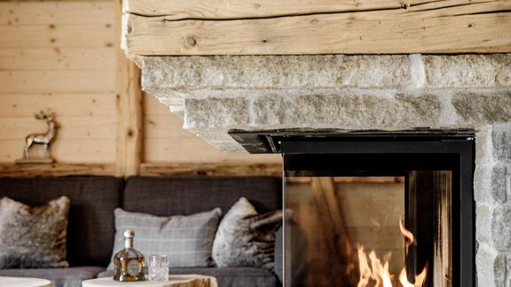 A cozy fireplace with a blazing fire and rustic stone frame. Decorative deer figures stand on the mantel, while a comfortable seating area is located in the background. - LENI MOUNTAIN CHALET - Sölden