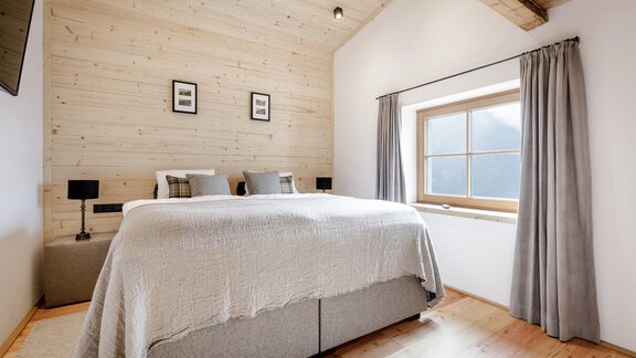 A modern bedroom with a large bed and wooden decor. The room has a window with curtains and bright light. - LENI MOUNTAIN CHALET - Sölden