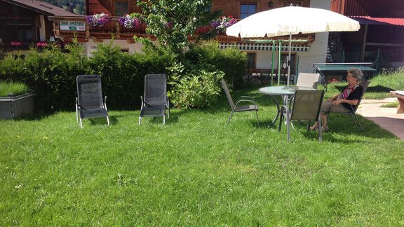 A sunny garden with green grass and several lounge chairs. A person is sitting relaxed at a table under a sun umbrella. - Pension Birkenheim - Sölden