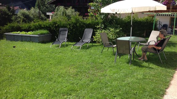 A quiet garden with green grass, a table and chairs under a parasol. In the background, deck chairs and a raised bed are visible. - Pension Birkenheim - Sölden
