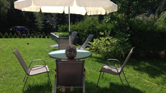 A beautiful garden with a round table and four chairs. A parasol provides shade while someone sits and enjoys the surroundings. - Pension Birkenheim - Sölden