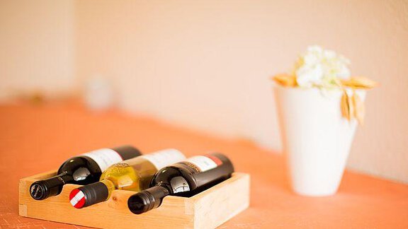 Three bottles of wine in a wooden rack are sitting on a table. Next to the wine is a vase with a decorative flower. - Pension Charly - Sölden