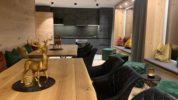 An elegant room with a paneled wooden wall and a modern kitchen. In the center is a large wooden table with a golden decorative reindeer and comfortable, stylish chairs. - Pension Sportalm - Sölden