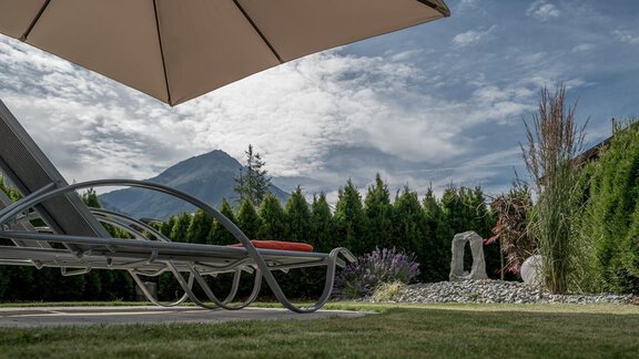 A relaxing lounger in the garden under a sun umbrella. In the background, a few trees and a mountain landscape can be seen. - Residenz Allegra - Sölden | © Residenz Allegra Sölden