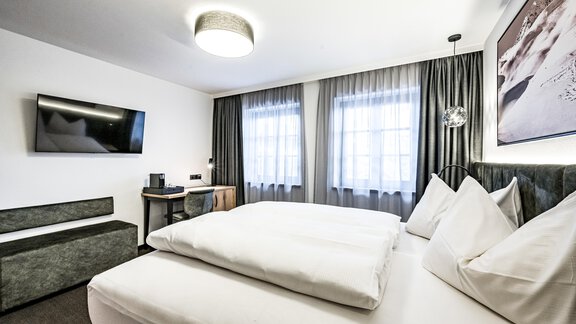 A modern hotel room with a large double bed and two pillows. There is a flat-screen TV and large windows with curtains. - Sonnalp Residences - Sölden
