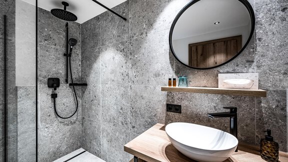 A modern bathroom with a glass shower and gray stone walls. On the wooden shelf, there is a sink and a large round mirror. - Sonnalp Residences - Sölden