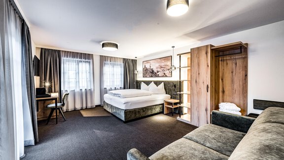 A modern hotel room with a large bed, a sofa, and a desk. Bright windows provide plenty of light and a pleasant atmosphere. - Sonnalp Residences - Sölden