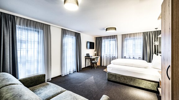 A modern hotel room with a large bed, a small workspace, and a cozy sofa.  
Large windows with curtains create a bright and inviting space. - Sonnalp Residences - Sölden
