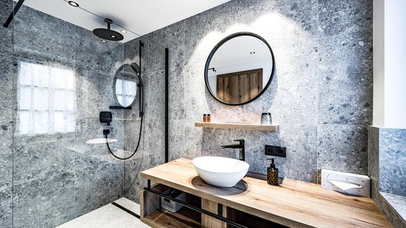 A modern bathroom with gray stone walls and an elegant shower. A ceramic sink sits on a wooden shelf. - Sonnalp Residences - Sölden