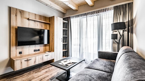 A modern living room with a gray sofa and a television on the wall. Large windows let in plenty of light and create a bright atmosphere. - Sonnalp Residences - Sölden