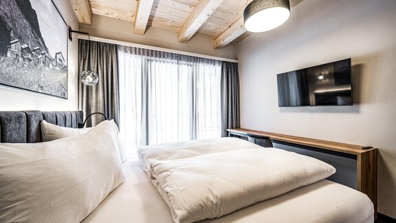 A modern bedroom with a large bed and wooden ceilings. Large windows let in plenty of light, and there is a television on the wall. - Sonnalp Residences - Sölden
