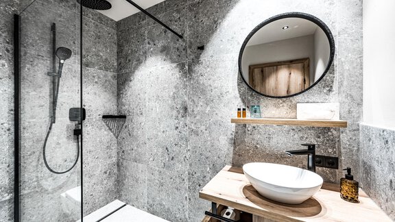 A modern bathroom with a glass shower and a stylish wall made of gray stone. On a wooden table, there is a sink and decorative care products. - Sonnalp Residences - Sölden