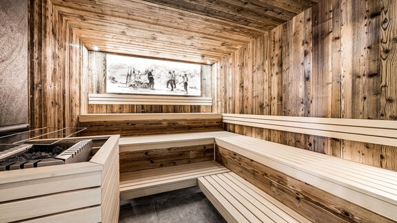 A modern sauna with wooden paneling and spacious benches. The room has a relaxing atmosphere with a large window and a piece of artwork on the wall. - Sonnalp Residences - Sölden