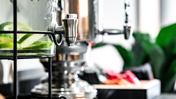 An elegant beverage dispenser with fresh herbs and citrus fruits is placed on a modern counter. In the background, plants and a stylish decor are visible. - Sonnalp Residences - Sölden
