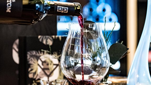 An elegant glass is filled with red wine, while a small shot stands beside it. In the background, stylish decorations and a wine storage are visible. - Sonnalp Residences - Sölden