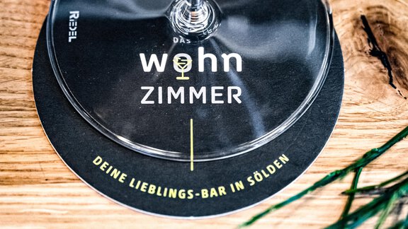A glass of wine stands on a wooden surface. Next to it lies a round coaster with the inscription "living ROOM". - Sonnalp Residences - Sölden