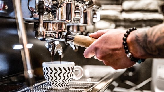A hand holds a tamper and prepares fresh espresso in a cup. In the background, a modern coffee machine is visible. - Sonnalp Residences - Sölden