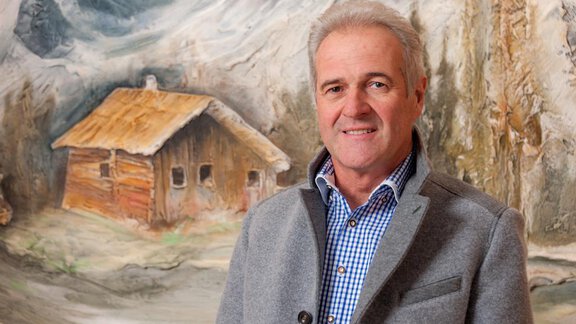 A man stands in front of a picturesque landscape with a wooden house. In the background, mountains and trees are visible. - Top Apart Daniela - Sölden