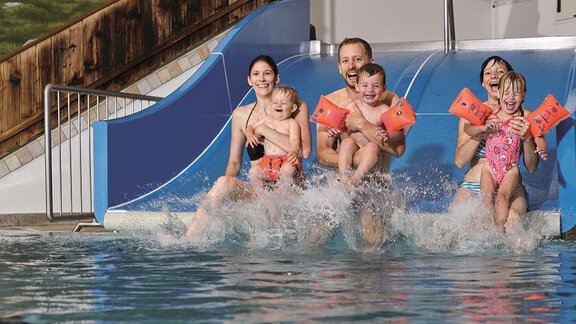 A cheerful family slides down a waterslide into the swimming pool together. Everyone is having fun and wearing swim floaties. - Top Apart Daniela - Sölden