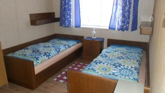 A simple bedroom with two beds, a lamp, and a small nightstand. Blue curtains hang in front of the window. - Appt. Garni Anny - Vent
