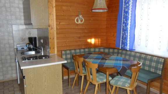 A cozy kitchen with wooden paneling and a dining table. In the background, there are windows with curtains and a small kitchenette. - Appt. Garni Anny - Vent