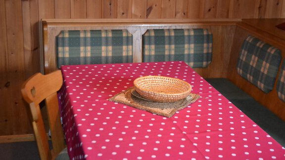 An inviting dining area with a round table decorated with a red-and-white tablecloth. The walls are made of wood, and the seats are upholstered with checked fabric. - Appt. Garni Anny - Vent