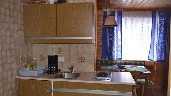 A small kitchen with wooden furniture and a sink. Next to the kitchenette is a table with chairs and a window with blue curtains. - Appt. Garni Anny - Vent