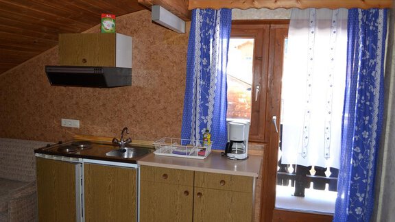 A small kitchen unit with wooden shelves and a window that lets in plenty of light. Curtains in blue and white complement the cozy space. - Appt. Garni Anny - Vent