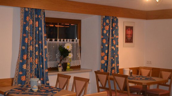 A cozy dining area with wooden chairs and tables. The windows are adorned with blue curtains and a floral decoration. - Appt. Garni Anny - Vent