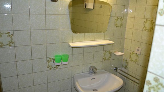 A small bathroom with green tiled walls. There is a sink, a mirror, and shelves. - Appt. Garni Anny - Vent