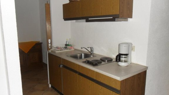 A small, functional kitchen unit with wooden cabinets and a sink. It includes cooking areas and a coffee machine. - Appt. Garni Anny - Vent