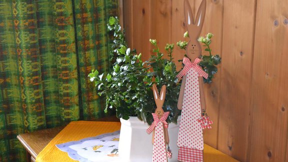 A decorative table with a white flower pot and green plants. Next to it stand two wooden Easter bunnies with red bows. - Appt. Garni Anny - Vent