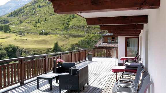 A large terrace with wood and comfortable furniture. In the background, green hills and mountains can be seen. - Gästehaus Soldanella - Vent