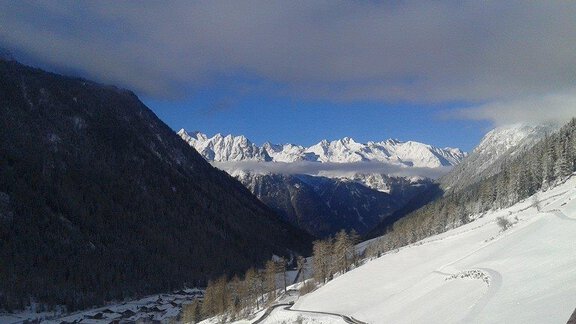 A winter landscape with snow-covered mountains and a blue sky. The valley is visible with snow-covered trees and a small village. - Berghof Schöpf - Gries