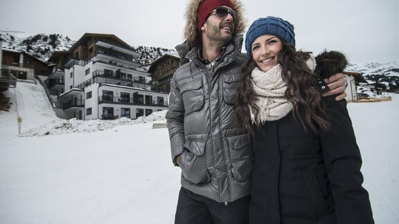 A couple stands in a snow-covered area, smiling and dressed in warm winter clothing. In the background, modern chalets and snow-capped mountains can be seen. - Gletscherblick - Serviced Apartments - Obergurgl 