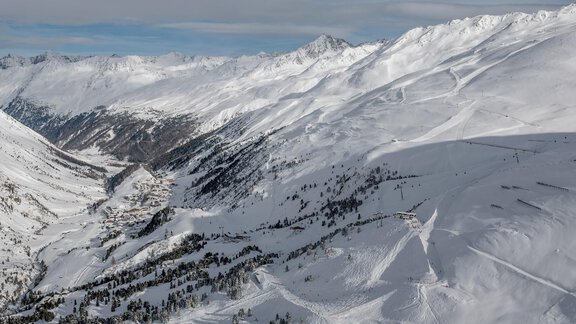 A picturesque winter landscape with snow-covered mountains and a clear sky. In the valley, scattered trees are visible, contrasting with the white snow. - Gletscherblick - Serviced Apartments - Obergurgl 