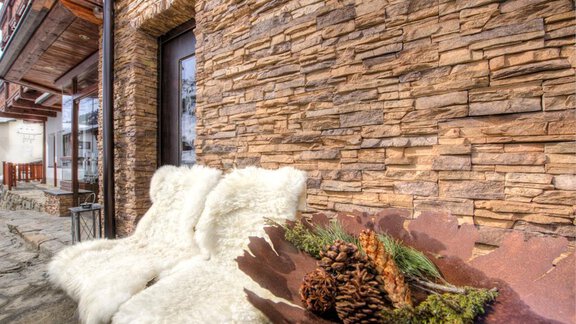 A cozy seating area with white furs in front of a stone wall. On a rustic tray, there are pine cones and green branches. - Hotel-Garni Pirchhütt - Obergurgl 