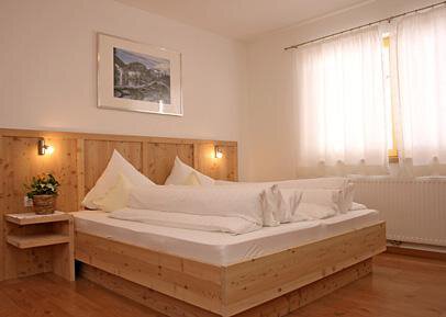 A cozy bedroom with a large double bed and wooden details. The room is bright and cheerfully lit by a window with curtains. - Pension Angern - Obergurgl 