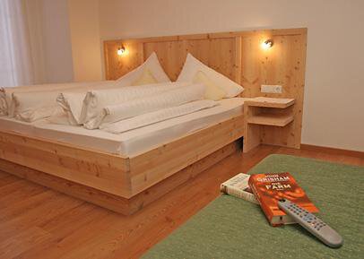 A modern bedroom with a wooden bed and white cushions. On the nightstand lies a book and a remote control. - Pension Angern - Obergurgl 