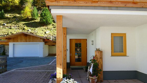 A modern wooden house with an inviting entrance. In the foreground, plants are blooming and in the background, other buildings and trees are visible. - Pension Angern - Obergurgl 