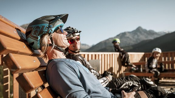 Eine Gruppe von Mountainbikern entspannt sich auf einer Holzbank mit Blick auf die Berge. Die Sonne scheint und die Atmosphäre ist entspannt. - Gasthof Waldesruh - Ochsengarten