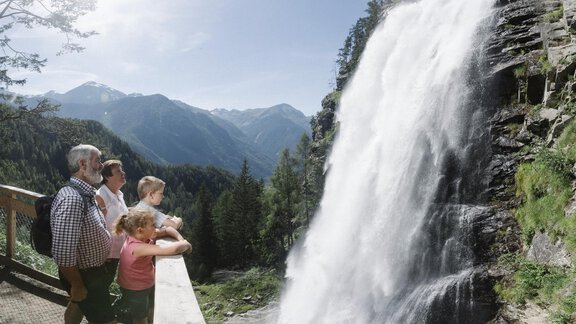 Eine Familie steht an einem Aussichtspunkt und betrachtet einen beeindruckenden Wasserfall in den Bergen. Die Sonne scheint und die Umgebung ist grün und naturnah. - Gasthof Waldesruh - Ochsengarten