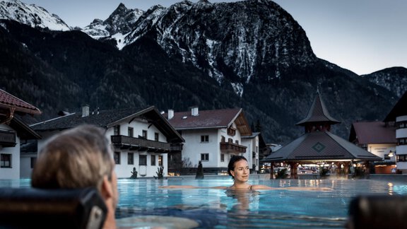 Ein entspannender Pool mit Blick auf majestätische Berge. Im Hintergrund sind traditionelle Chalets und eine stimmungsvolle Abendbeleuchtung zu sehen. - Posthotel Kassl - Oetz