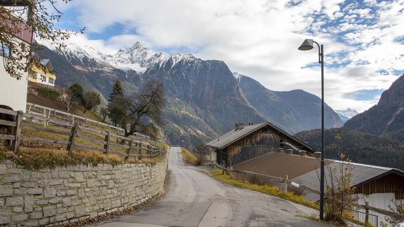 Eine ruhige Straße in den Bergen mit schneebedeckten Gipfeln im Hintergrund. Der Weg ist gesäumt von traditionellen Häusern und einem Steinzaun. - Sternenvilla - Oetz | © Markus Maas