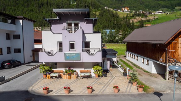 Ein modernes Gebäude in lila mit einem gepflegten Eingangsbereich und Blumenkübeln. Im Hintergrund sind grüne Berge zu sehen. - Alpenapart Saphir - Sölden