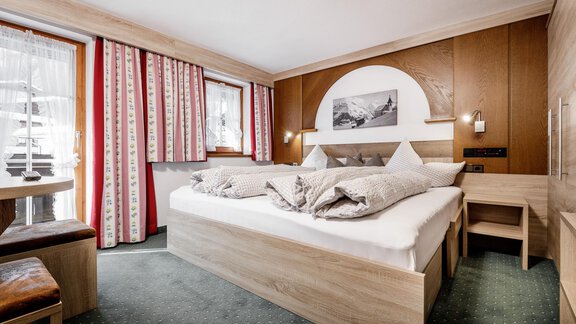 A cozy hotel room with a large bed and modern wooden furniture. The room features large windows with curtains and an inviting atmosphere. - Altes Holzhaus - Sölden