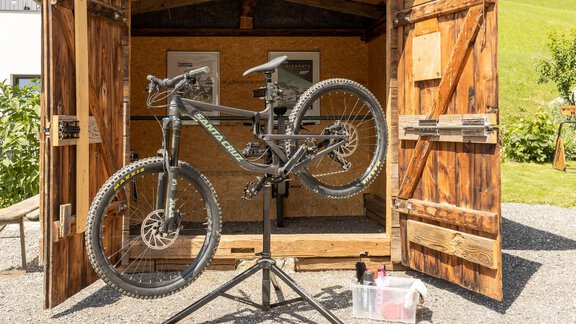 A bicycle is on a maintenance stand in an open wooden shed. In the background, green meadows and a beautiful sky can be seen. - Altes Holzhaus - Sölden