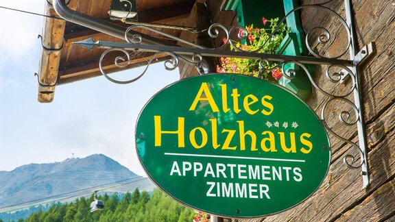 A sign of the old wooden house with the inscription "Old Wooden House – Apartments Rooms". In the background, mountains and trees are visible. - Altes Holzhaus - Sölden