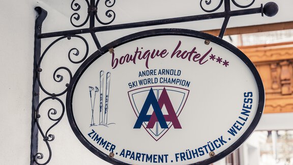 A hanging sign for a boutique hotel. It displays the name of the hotel and some services like rooms, apartment, and wellness. - Andre Arnold - Boutique Hotel - Sölden