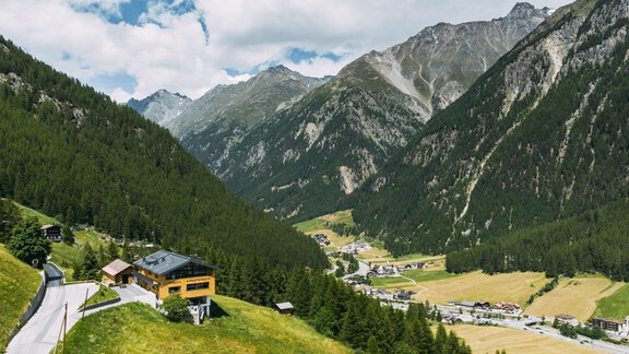 A breathtaking mountain landscape with green meadows and high mountains. In the foreground stands a modern house, while the valley is visible in the distance. - Anraitl Appartements Sölden - Sölden
